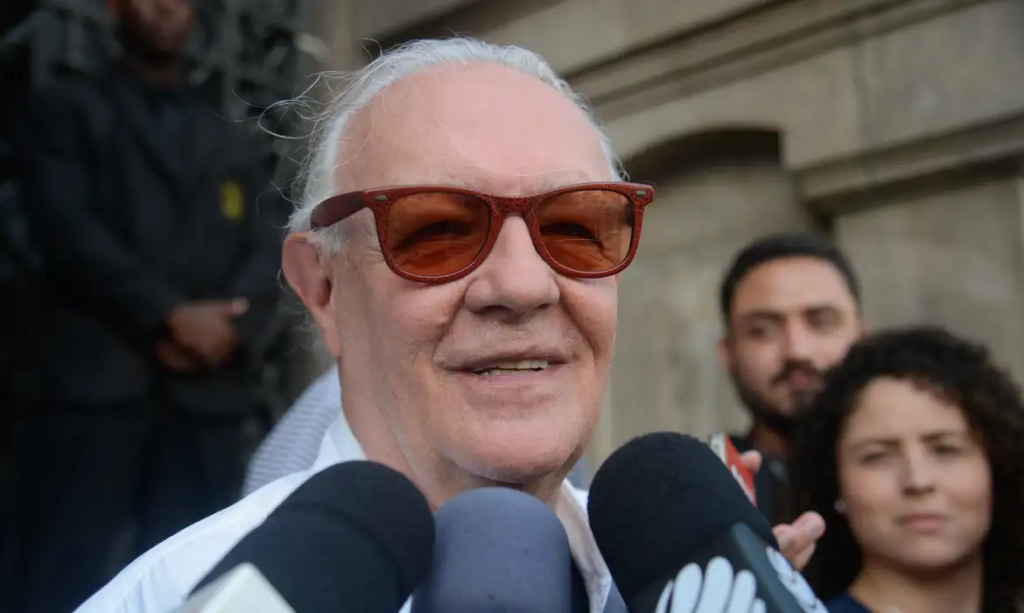 Ney Latorraca sorrindo durante entrevista, usando óculos de sol, com microfones à sua frente.