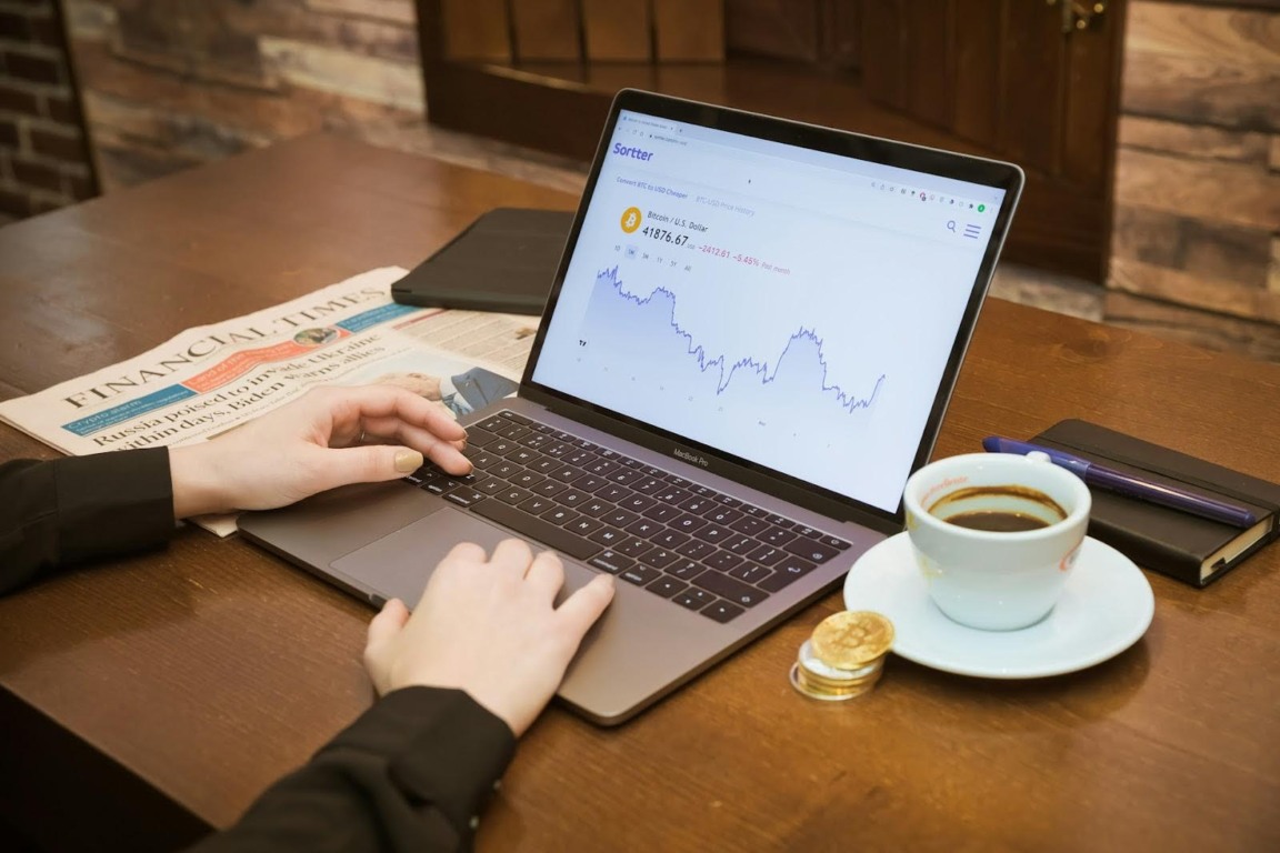 Pessoa usando notebook com gráfico de criptomoeda na tela, ao lado de café, moedas douradas e jornal, representando o mercado digital e financeiro.