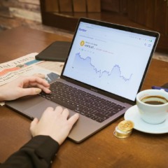 Pessoa usando notebook com gráfico de criptomoeda na tela, ao lado de café, moedas douradas e jornal, representando o mercado digital e financeiro.