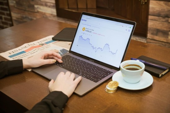 Pessoa usando notebook com gráfico de criptomoeda na tela, ao lado de café, moedas douradas e jornal, representando o mercado digital e financeiro.