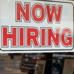 Placa "Now Hiring" em janela de loja nos EUA, refletindo a importância do mercado de trabalho americano para o calendário econômico e o payroll.