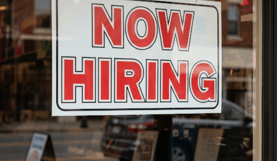 Placa "Now Hiring" em janela de loja nos EUA, refletindo a importância do mercado de trabalho americano para o calendário econômico e o payroll.