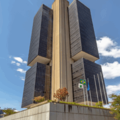 Prédio do Banco Central do Brasil em Brasília em um dia ensolarado, visto de baixo, com céu azul e árvores ao redor.