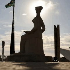 Vista frontal da estátua "A Justiça" em silhueta contra um sol brilhante. A estátua representa uma figura feminina sentada, vendada, segurando uma espada. À esquerda, um mastro alto com a bandeira do Brasil. Ao fundo, parte da arquitetura moderna do prédio do STF e um céu com algumas nuvens.