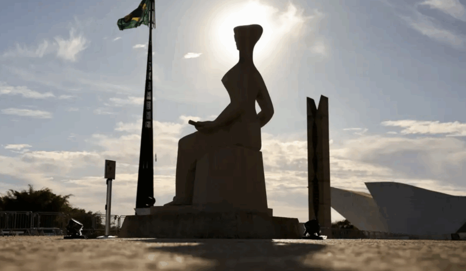 Vista frontal da estátua "A Justiça" em silhueta contra um sol brilhante. A estátua representa uma figura feminina sentada, vendada, segurando uma espada. À esquerda, um mastro alto com a bandeira do Brasil. Ao fundo, parte da arquitetura moderna do prédio do STF e um céu com algumas nuvens. Vista frontal da estátua "A Justiça" em silhueta contra um sol brilhante. A estátua representa uma figura feminina sentada, vendada, segurando uma espada. À esquerda, um mastro alto com a bandeira do Brasil. Ao fundo, parte da arquitetura moderna do prédio do STF e um céu com algumas nuvens.
