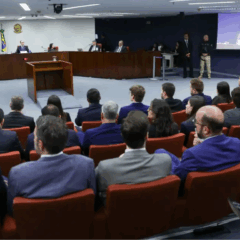 Pessoas sentadas em um auditório acompanhando um julgamento, com o painel de jurados à frente e uma tela exibindo o rosto de um homem falando.