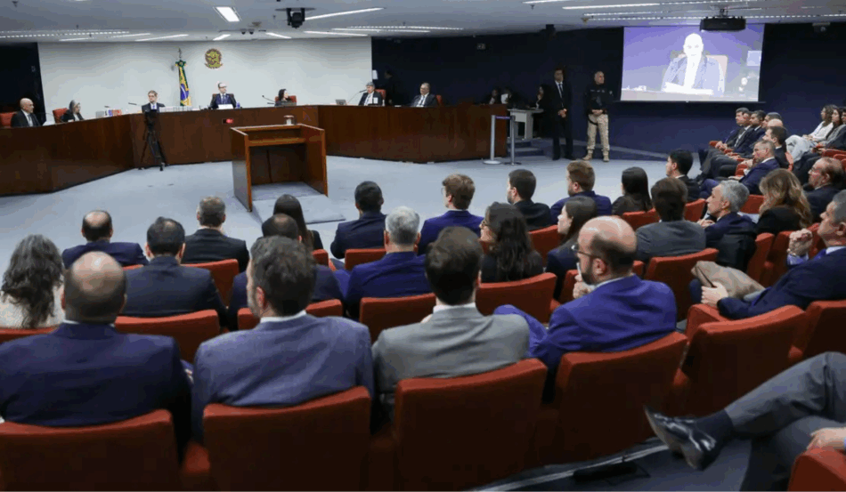 Pessoas sentadas em um auditório acompanhando um julgamento, com o painel de jurados à frente e uma tela exibindo o rosto de um homem falando. Pessoas sentadas em um auditório acompanhando um julgamento, com o painel de jurados à frente e uma tela exibindo o rosto de um homem falando.