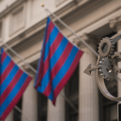 Detalhe de um poste com símbolo circular de engrenagens e setas, em primeiro plano, com bandeiras listradas de azul e vermelho e um edifício clássico em segundo plano