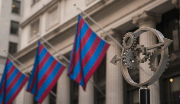 Detalhe de um poste com símbolo circular de engrenagens e setas, em primeiro plano, com bandeiras listradas de azul e vermelho e um edifício clássico em segundo plano