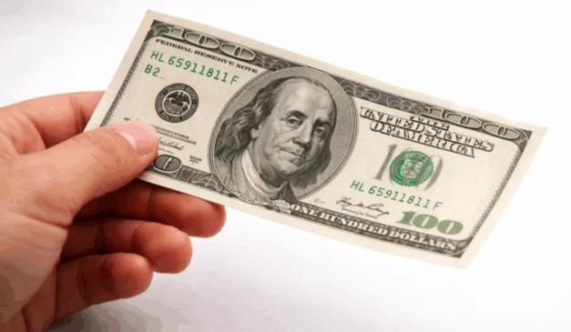 Mão masculina segurando uma nota de 100 dólares, representando a queda do dólar frente ao real no encerramento do ano