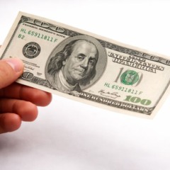 M&atilde;o masculina segurando uma nota de 100 d&oacute;lares, representando a queda do d&oacute;lar frente ao real no encerramento do ano