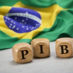 Pe&ccedil;as de madeira com a sigla PIB sobre superf&iacute;cie, com bandeira do Brasil desfocada ao fundo, representando a economia brasileira