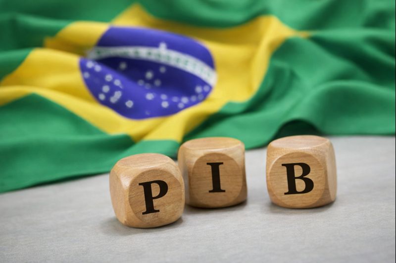 Peças de madeira com a sigla PIB sobre superfície, com bandeira do Brasil desfocada ao fundo, representando a economia brasileira